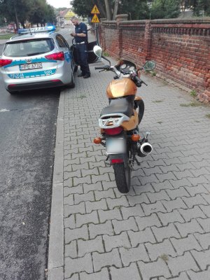 Na pierwszym planie widoczny motorower, na drugim planie stoi radiowóz, obok niego policjant