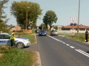 Na zdjęciu ukazany jest policjant stojący na poboczu obok radiowozu obserwujący przejazd kolejowy