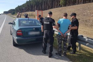 Na zdjęciu widoczna jest samochód zatrzymanego. Zatrzymany stoi obok samochodu i jest pilnowany przez trzech policjantów. W tle za samochodem widoczni są operatorzy kamery nagrywający całą sytuację.