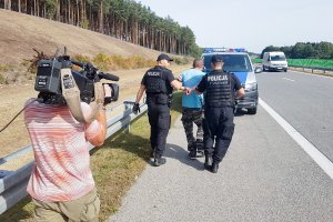 Zdjęcie przedstawia operatora kamery, który to nagrywa jak dwóch policjantów odprowadza zatrzymanego skutego w kajdankach do policyjnego busa.