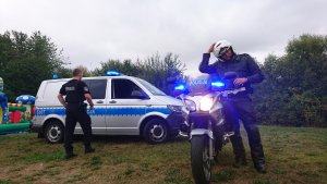 Na zdjęciu widoczni są dwaj policjanci, jeden z nich jest kierowcą motocykla. Na drugim planie jest bus policyjny oraz motocykl policyjny, w tle widać również kawałek dmuchanego zamku do zabawy dla dzieci.