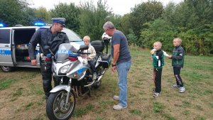 Na zdjęciu jest policjant, który pozwala usiąść na motocyklu dzieciom, które podeszły zainteresowane wraz z ich opiekunem. W tle widoczny jest także policyjny bus. W obydwu pojazdach są uruchomione sygnały świetlne.