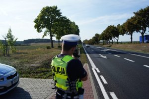 Na zdjęciu widnieje policjant stojący obok radiowozu przy drodze, trzymając w ręce fotoradar, dokonuje rutynowej kontroli prędkości na drodze nadjeżdzającego pojazdu z naprzeciwka.