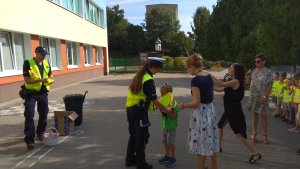 Zdjęcie przedstawia policjantów, którzy ubierają dziecku kamizelke odblaskową, w tle widoczne są inne dzieci oraz nauczycielki, które mówią do mikrofonu.