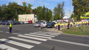 Zdjęcie przedstawia dzieci czekających przed przejściem dla pieszych wraz z policjantami. W tle widoczny jest również policjant kierujący ruchem na drodze.