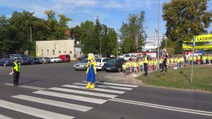 Zdjęcie przedstawia dzieci czekających przed przejściem dla pieszych wraz z policjantami. W tle widoczny jest również policjant kierujący ruchem na drodze, oraz człowiek maskotka przechodzący przez przejście dla pieszych.