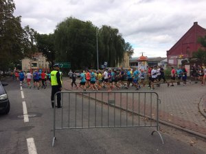 Na zdjęciu widzimy ludzi biegnących w biegu. W tle widoczna jest także policjant, który zabezpiecza dany bieg.