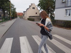 Na zdjęciu widzimy młodą kobietę, która przechodząc przez przejście dla pieszych, używa telefonu komórkowego, jednocześnie nie zwracając żadnej swojej uwagi na otoczenie.