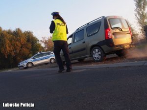 Zdjęcie przedstawia policjantkę, która podchodzi do kontrolowanego przez nią pojazdu. W tle stoi także radiowóz.