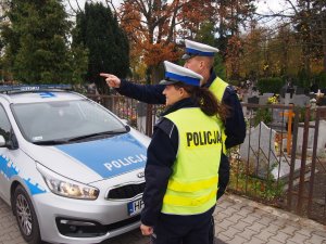 Na zdjęciu widać policjantów rozmawiających ze sobą obok radiowozu. W tle widoczny jest cmentarz.