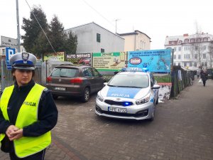 Na zdjęciu widoczna jest policjantka stojąca obok radiowozu stojącego na parkingu