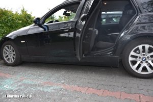 Czarne auto osobowe z otwartymi tylnymi drzwiami i widocznym fotelikiem. Przednie drzwi od kierowcy nie mają szyby a na bruku leży rozbite szkło.