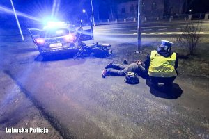 osoba zatrzymana leżąca na ziemi, przy niej funkcjonariusz policji, w tle radiowóz z włączonymi sygnałami świetlnymi