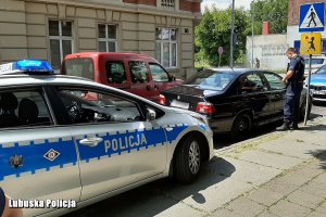 stojący radiowóz, a przed nim samochód zatrzymanego marki BMW, przy którym stoi policjant