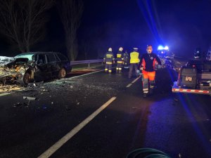 Idący drogą ratownik medyczny, w tle dwóch strażaków oraz policjant, po ich bokach dwa rozbite auta, noc