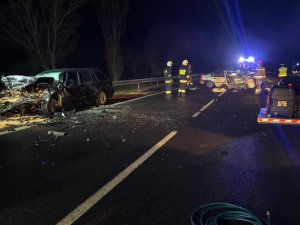 Na drodze stoją dwa rozbite auta, jest noc, widać troje strażaków, policjanta oraz jeden z pojazdów ma włączone szperacze