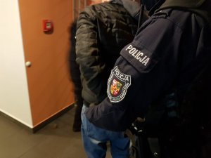 Dwóch policjantów prowadzi korytarzem mężczyznę, w tle widać kraty przegradzające korytarz. Postaciom widać sylwetki od ramion w dół. Na ramieniu policjanta widoczna jest naszywka z napisem Komenda Powiatowa Policji w Świebodzinie