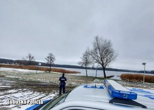 Policyjny radiowóz, a w tle policjant sprawdza czy nie ma żadnych osób na zamarzniętym jeziorze.