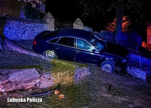 Na wzniesieniu widać uszkodzony kawałek kamiennego murku, powyżej postawione bokiem auto, którego przód, będący z prawej strony zawieszony jest na barierce przy schodach, w tle kościół