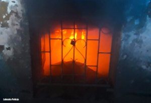 Okno piwniczne w kamienicy, górna część odymiona, w tle w pomieszczeniu widać ogień.