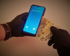 Dłonie w czarnych lateksowych rękawiczkach, w lewej ręce telefon na który dzwoni zastrzeżony numer a prawa z telefonu wyciąga dwa banknoty o nominale 200 i 100 zł