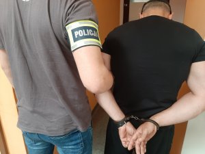 Mężczyzna stoi w asyście nieumundurowanego policjanta z opaską policja na ręce. Zatrzymany ma założone kajdanki na ręce z tyłu