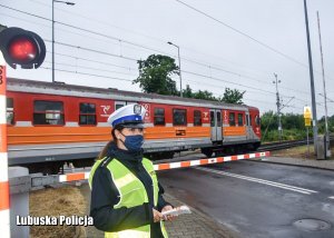 Policjantka z ruchu drogowego na tle przejazdu kolejowego. Przejeżdża pociąg