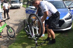 Na tle radiowozu policjant w stroju rowerzysty policyjnego pomaga dziecku przy rowerze.