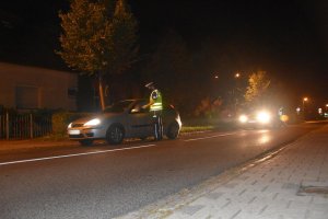 Umundurowany policjant oraz policjantka badają stan trzeźwości kierujących. Pojazdy stoją na ulicy oświetlonej lampami. Panuje zmrok