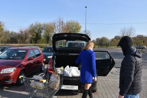Parking przy markecie, kobieta pakuje zakupy z wózka do bagażnika, część niesie na tylne siedzenie auta. W wózku stoi jej torebka.