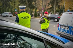 Policjant z Wydziału Ruchu Drogowego oraz funkcjonariusza Żandarmerii wojskowej, w tle cmentarz, w w pierwszym planie radiowozy.