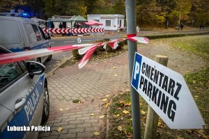 Widok na sklep i stragan ze zniczami, w pierwszym tle znak Cmentarz Parking i radiowozy oraz biało czerwona taśma.