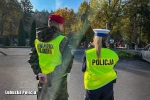 Policjantka z Wydziału Ruchu drogowego oraz Żandarm Wojska Polskiego wspólnie stoją w pobliżu cmentarza i pilnują porządku.