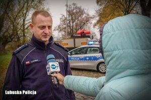 :Policjant na tle radiowozu, przed nim dziennikarz z mikrofonem.