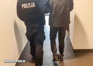 Korytarz, policjant prowadzi mężczyznę w kajdankach założonych na ręce i nogi