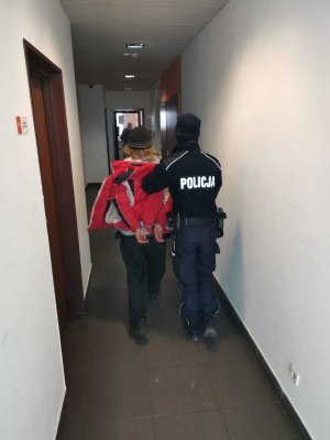 Korytarzem komendy, umundurowany policjant prowadzi zakutego w kajdanki mężczyznę.