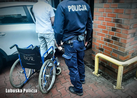 Policjant i sanitariusz pomagają wysiąść kobiecie na wózek, w tle brama wjazdowa na oddział ratunkowy szpitala