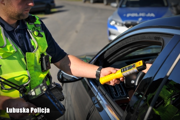 Policjant dokonuje kontroli z urządzeniem sprawdzającym trzeźwość, widok na rękę, urządzenie i okno auta od strony kierowcy