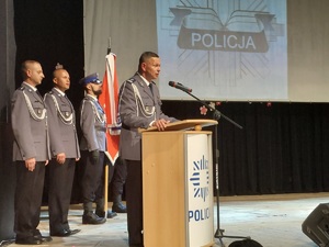 Komendant przemawia na scenie zza mównicy, z tyłu prowadzący uroczystość policjanci