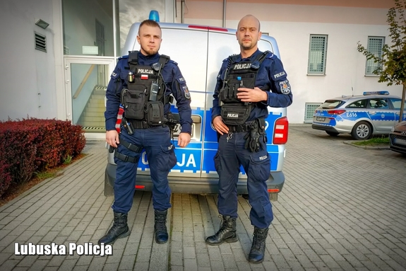 Dwóch umundurowanych policjantów na tle komendy i radiowozów.
