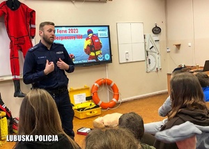 Klasa, plecy dzieci i w tle przemawiający policjant