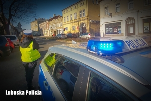 Radiowóz z sygnałami, w tle policjantka i przejście dla pieszych