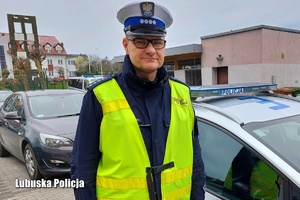 Policjant w umundurowaniu widziany od pasa w górę na tle radiowozu.