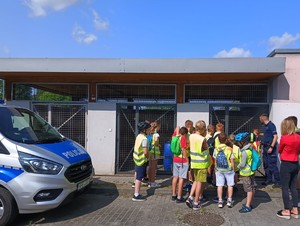 Policjant z grupą dzieci stojący przy policyjnych kojcach dla psów, z prawej strony widać część radiowozu.