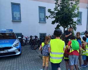 Policjant z dziećmi na policyjnym parkingu, widać radiowóz i rowery.