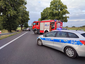 Ulica, na pierwszym planie radiowóz, wóz straży pożarnej, w tle auto po zderzeniu, na poboczu motocykl.