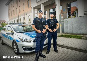 Dwóch policjantów przed radiowozem przy komendzie.