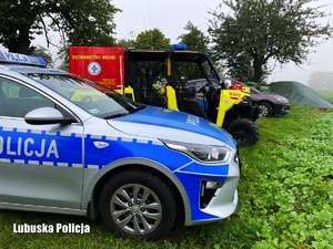 Pojazd policji, ratowników wodnych i cywilny na tle łąki i pól.