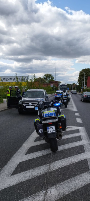 Radiowozy i motocykle na drodze, w tle pojazdy i policjanci obsługujący kolizję.