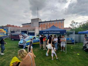 Policyjne stoisko, namiot, ludzie, mierzenie prędkości w biegu, przymierzanie strojów, dzieci uśmiechnięte w radiowozie.